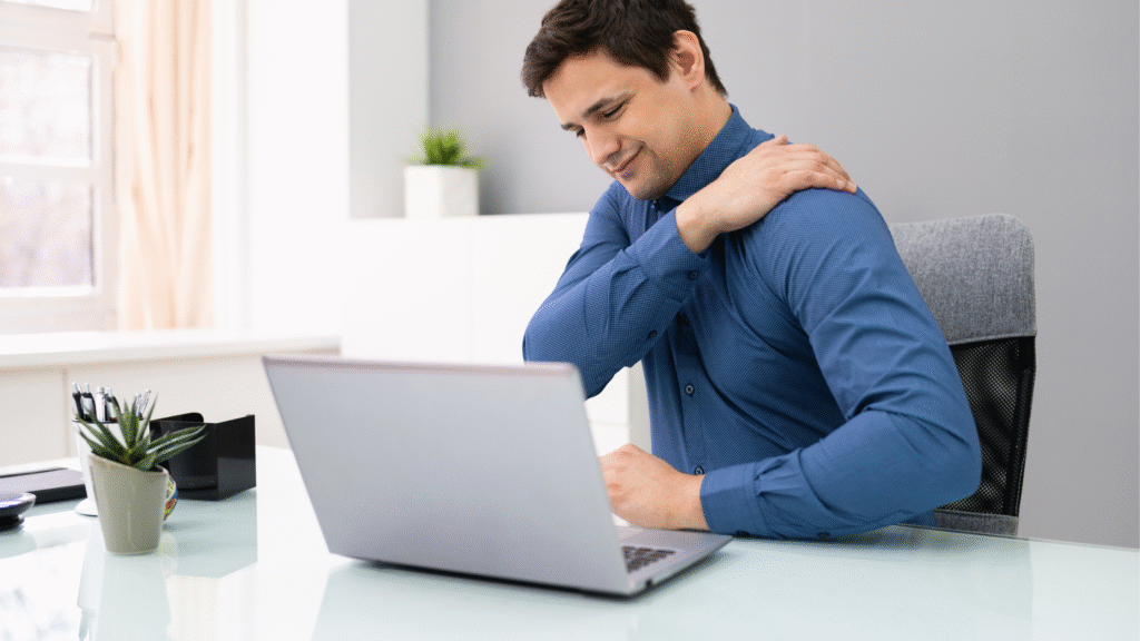 Man holding his shoulder in pain while working at a desk with a laptop.